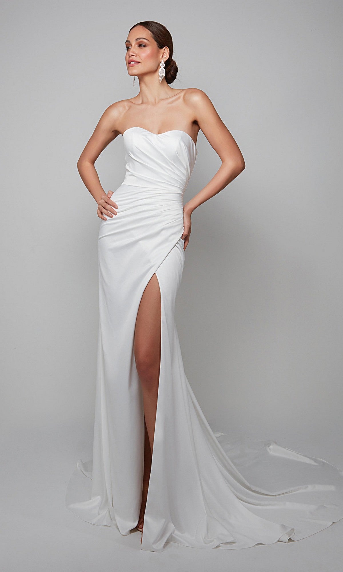 A woman poses confidently against a plain background, wearing the Alyce 7057 long white dress featuring a strapless design and high slit, with one hand on her hip.